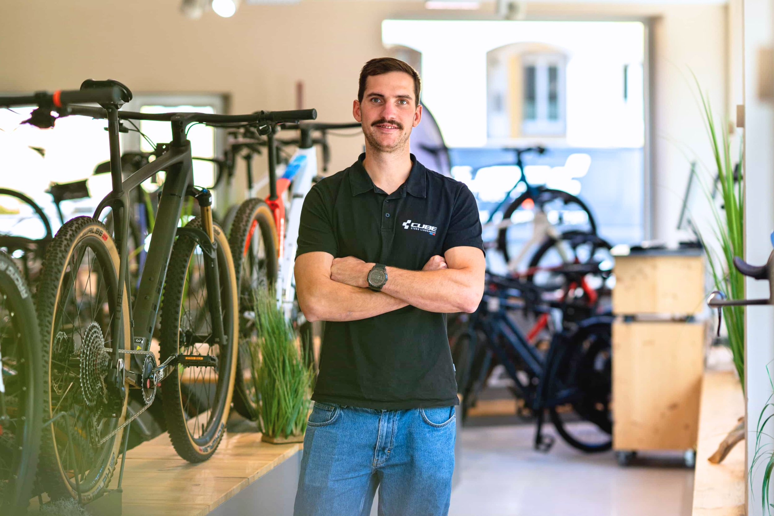 Ein Mann mit Schnurrbart steht lächelnd mit verschränkten Armen in einem Fahrradgeschäft, umgeben von verschiedenen ausgestellten Fahrrädern. Er trägt ein schwarzes Poloshirt und Jeans. Das Innere des Ladens ist hell erleuchtet.