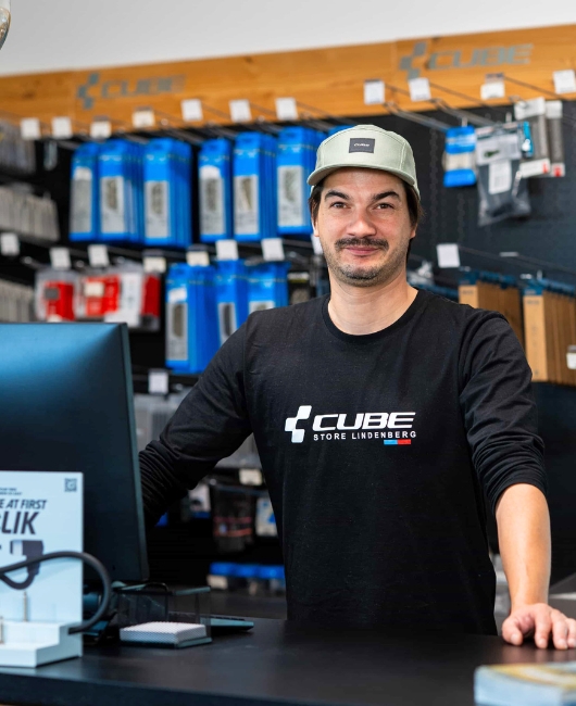 Ein Mann mit einer hellgrünen Mütze und einem schwarzen CUBE Store Lindenberg-Shirt steht lächelnd hinter einem Verkaufstresen. Hinter ihm sind die Regale mit Fahrradzubehör und -produkten aufgebaut.