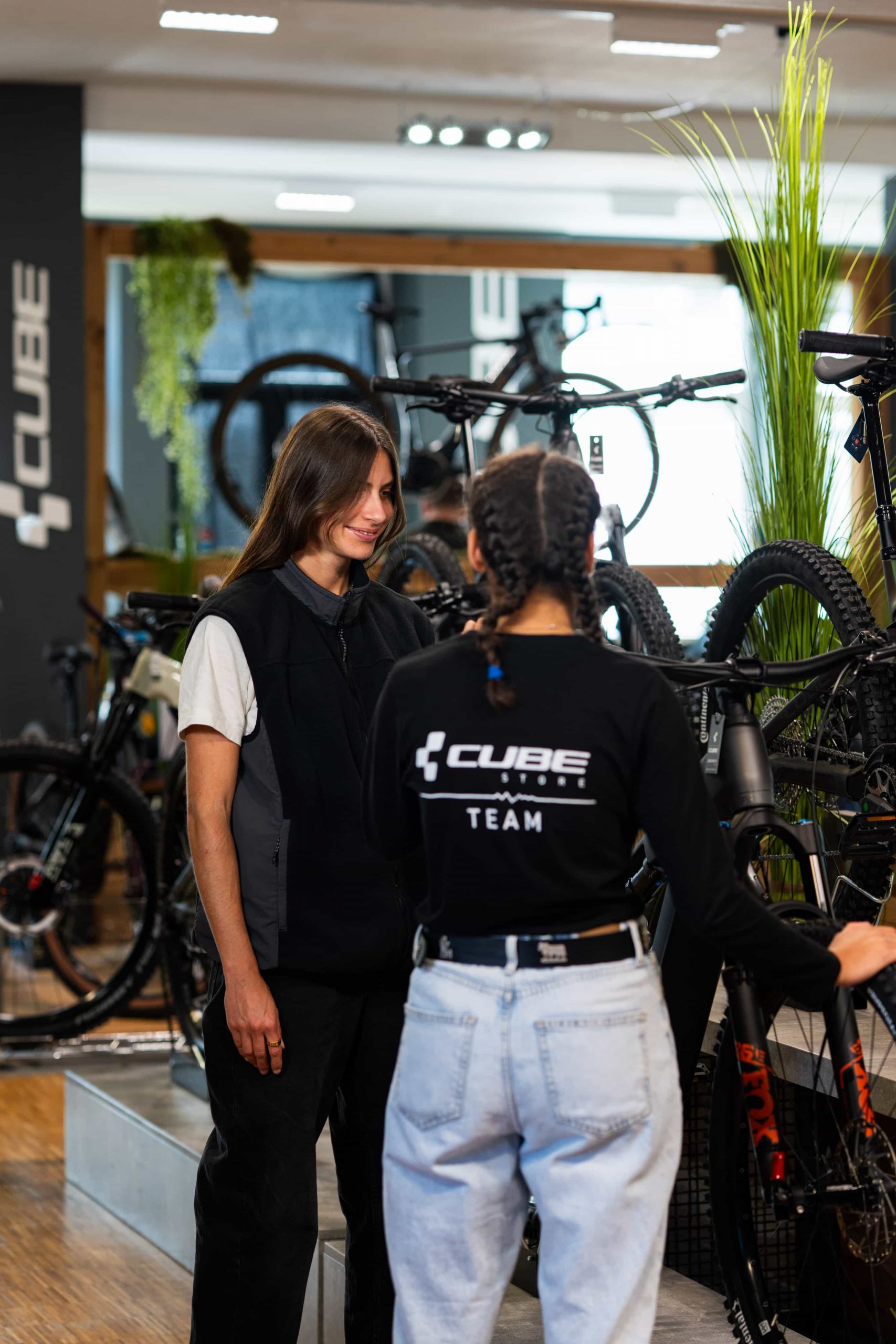 Zwei Frauen stehen in einem Fahrradgeschäft voller Mountainbikes und unterhalten sich. Die eine trägt ein CUBE TEAM-Shirt und weiße Jeans, die andere eine schwarze Weste. Fahrräder und Grünpflanzen schmücken den hellen, modernen Raum.