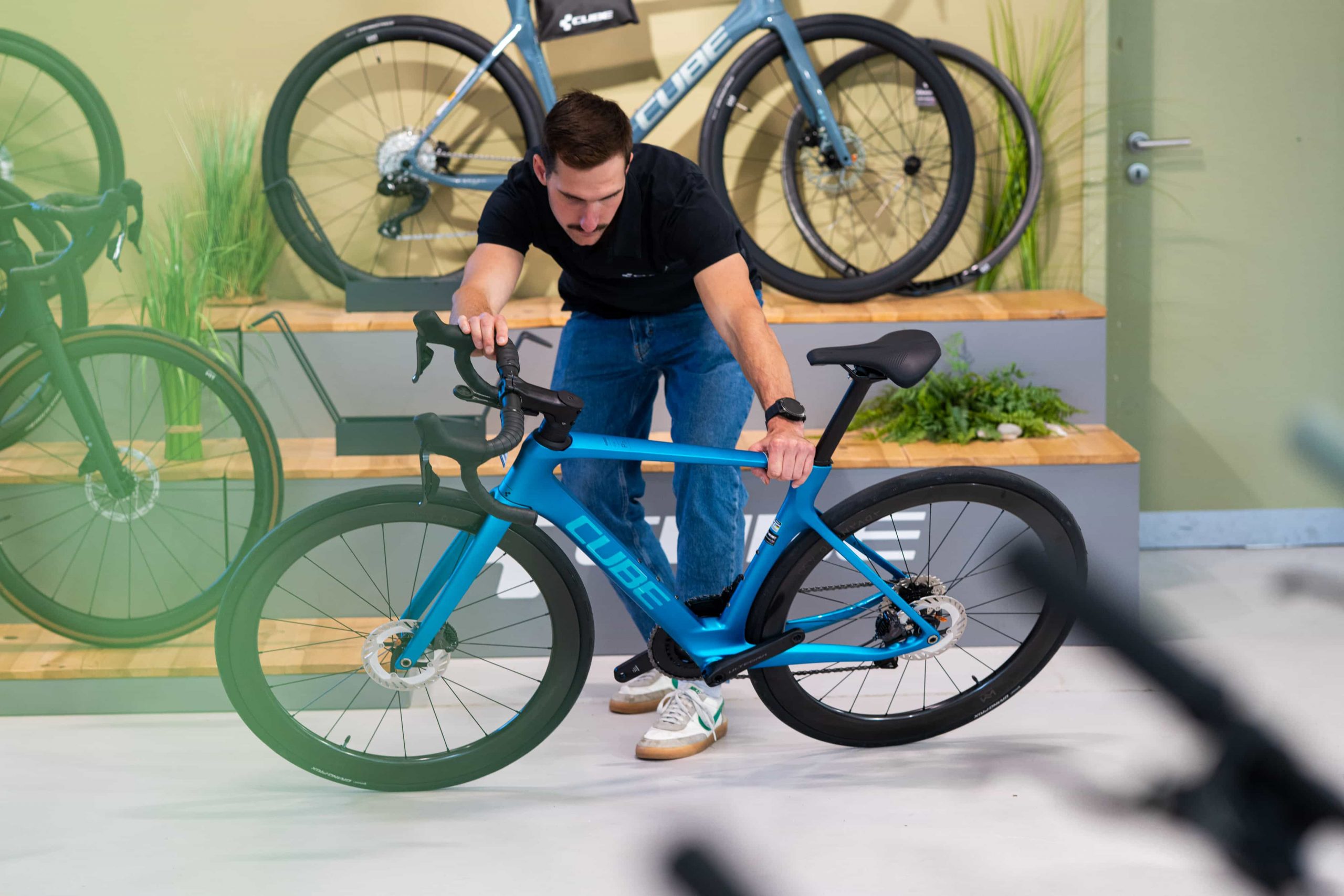 Ein Mann in schwarzem Hemd und Jeans beugt sich vor, um ein blaues Cube-Rennrad zu inspizieren oder einzustellen, während im Hintergrund weitere Fahrräder auf Holzplattformen ausgestellt sind.