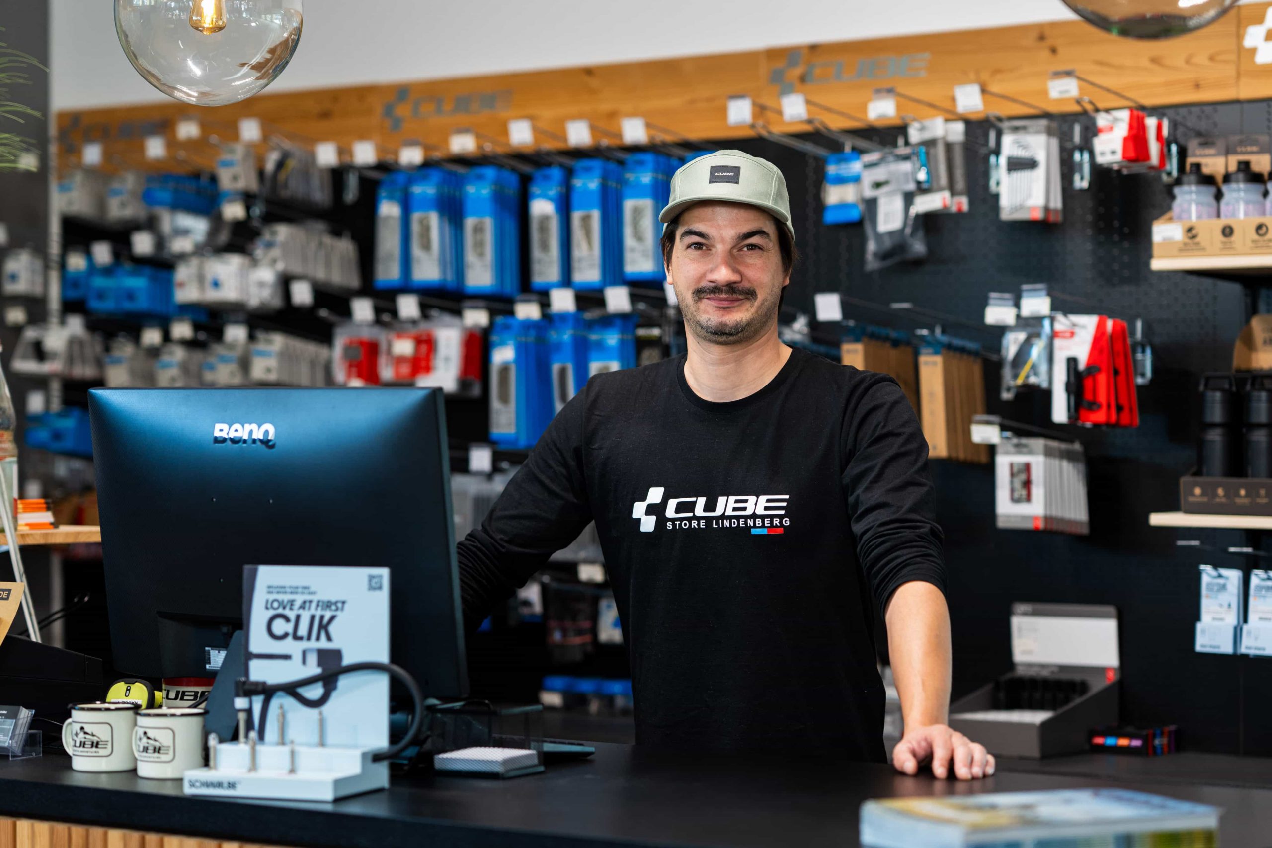 Ein Mann, der ein schwarzes CUBE-Shirt und eine Mütze trägt, steht lächelnd hinter der Theke eines Fahrradgeschäfts, an der Wand hinter ihm sind Fahrradzubehör und -produkte ausgestellt.