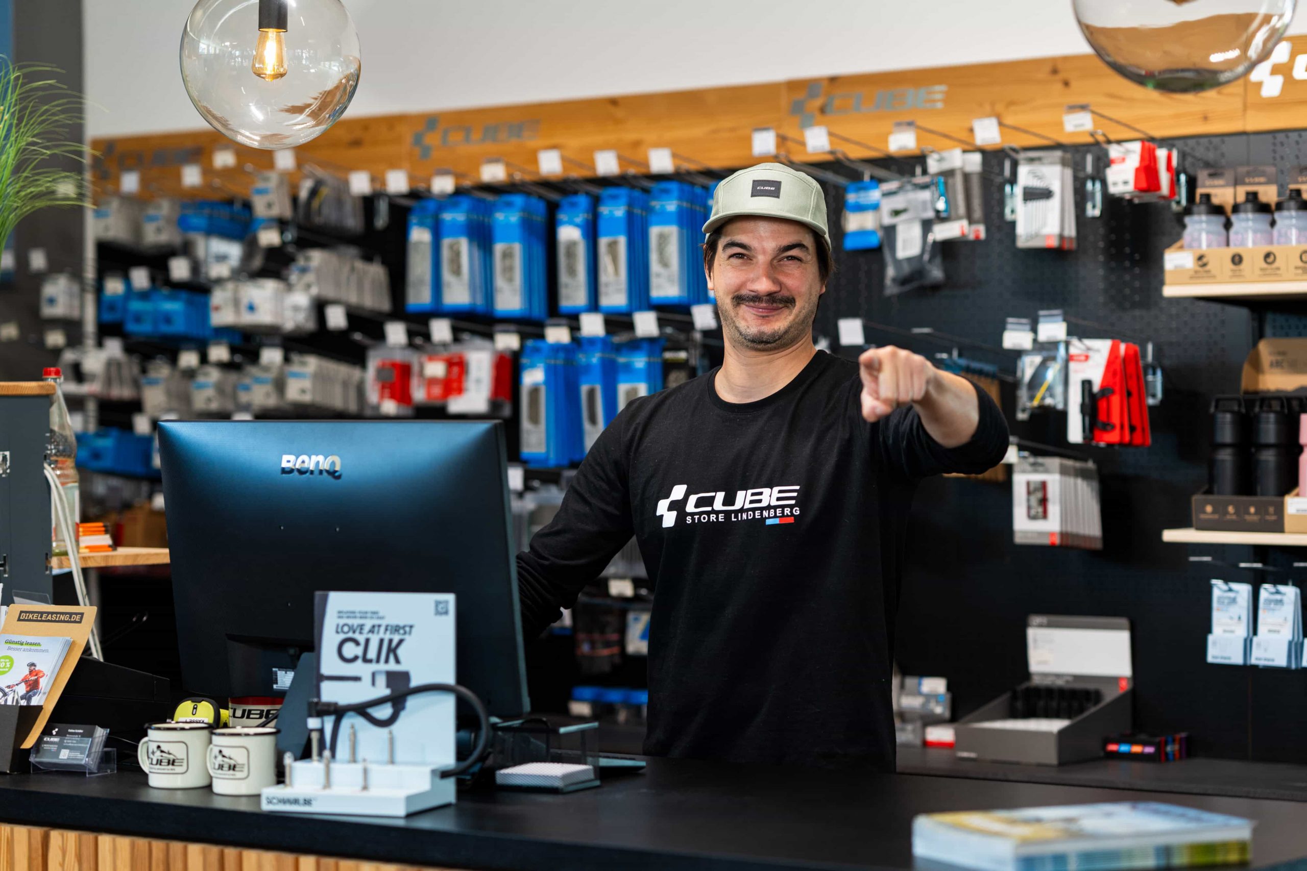 Ein lächelnder Angestellter mit Mütze und Cube-Shirt steht hinter dem Tresen eines Fahrradgeschäfts und zeigt in die Kamera. Auf dem Tresen stehen Tassen und ein Computer, an der Wand hinter ihm ist Fahrradzubehör ausgestellt.